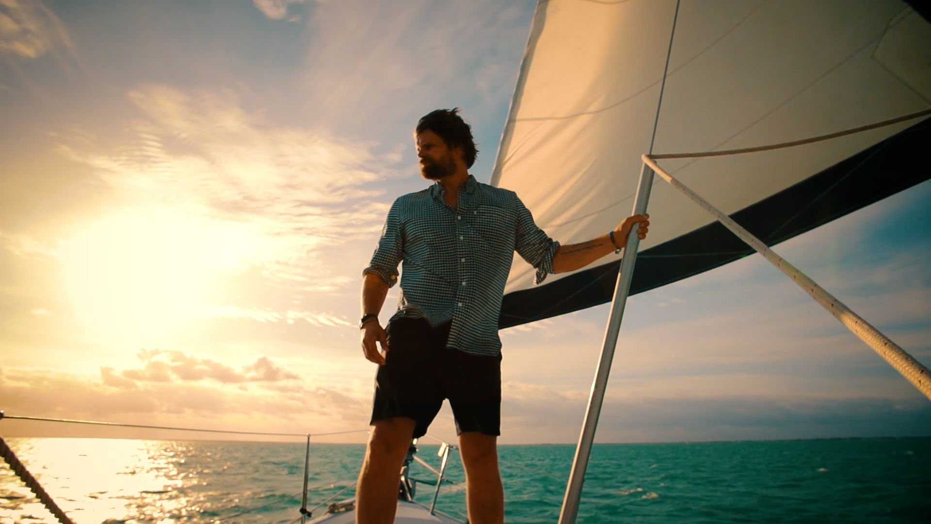 A man stands at the bow of a sailboat at sunset, holding the rigging.