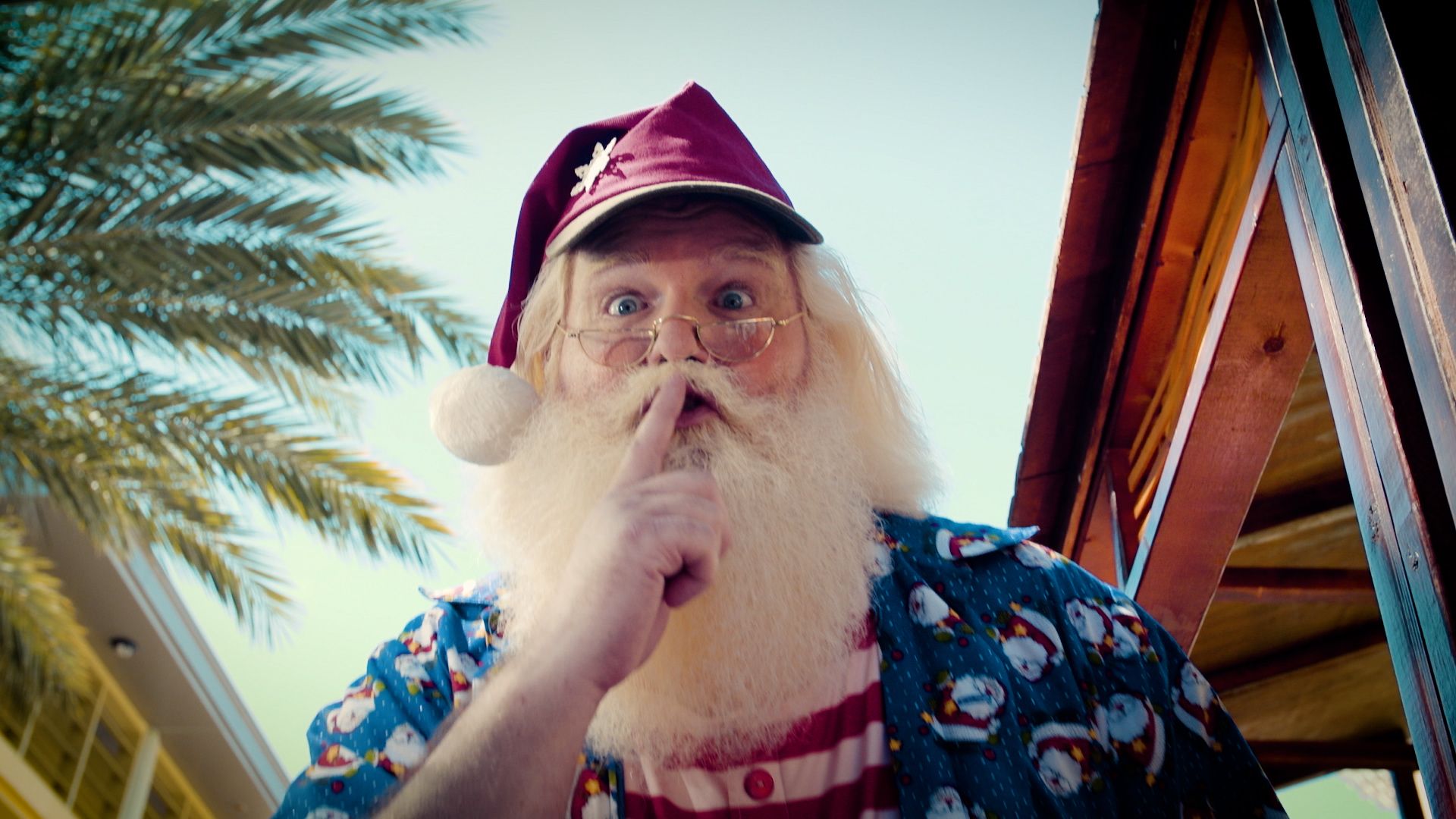Santa, in a cap and holiday shirt, holds a finger to his lips beneath a palm tree.