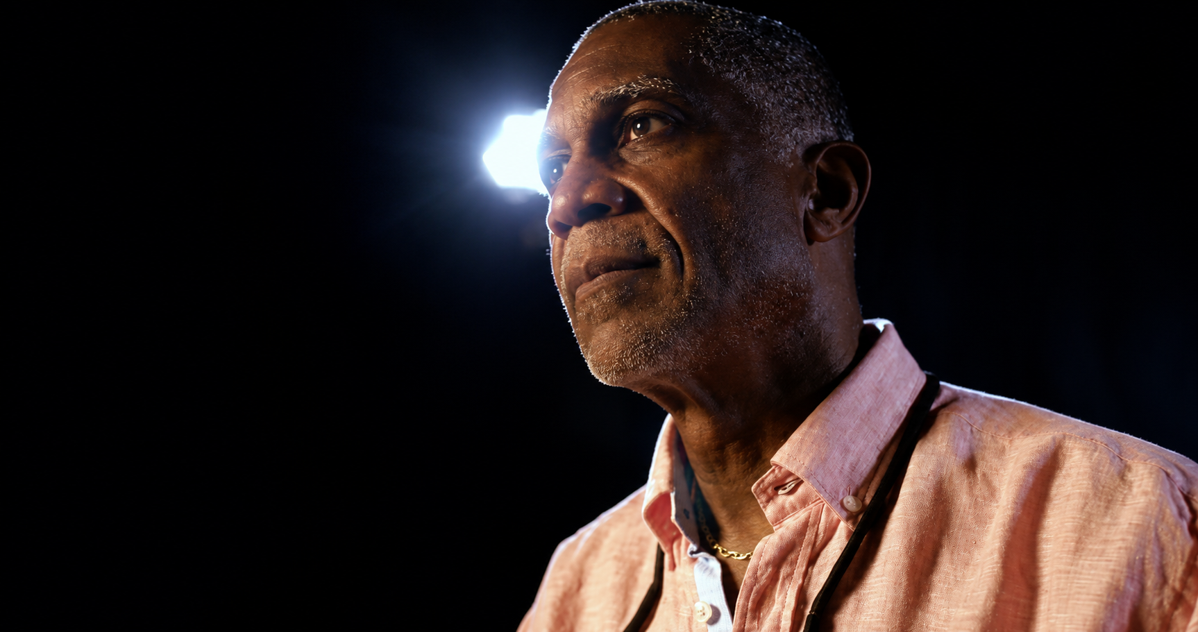 An older man in a pink shirt, lit by a single hard light against darkness.