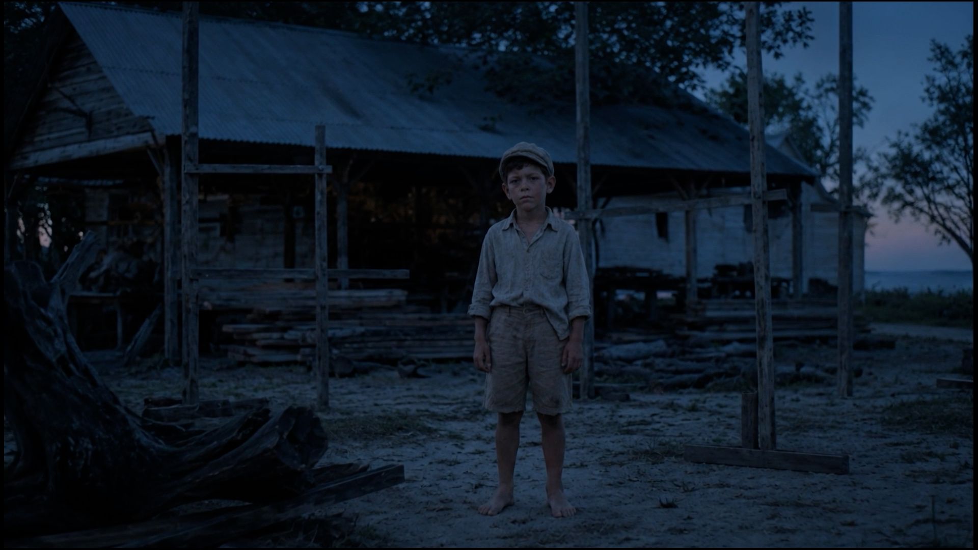 A boy stands barefoot in a dusk-lit boatyard.
