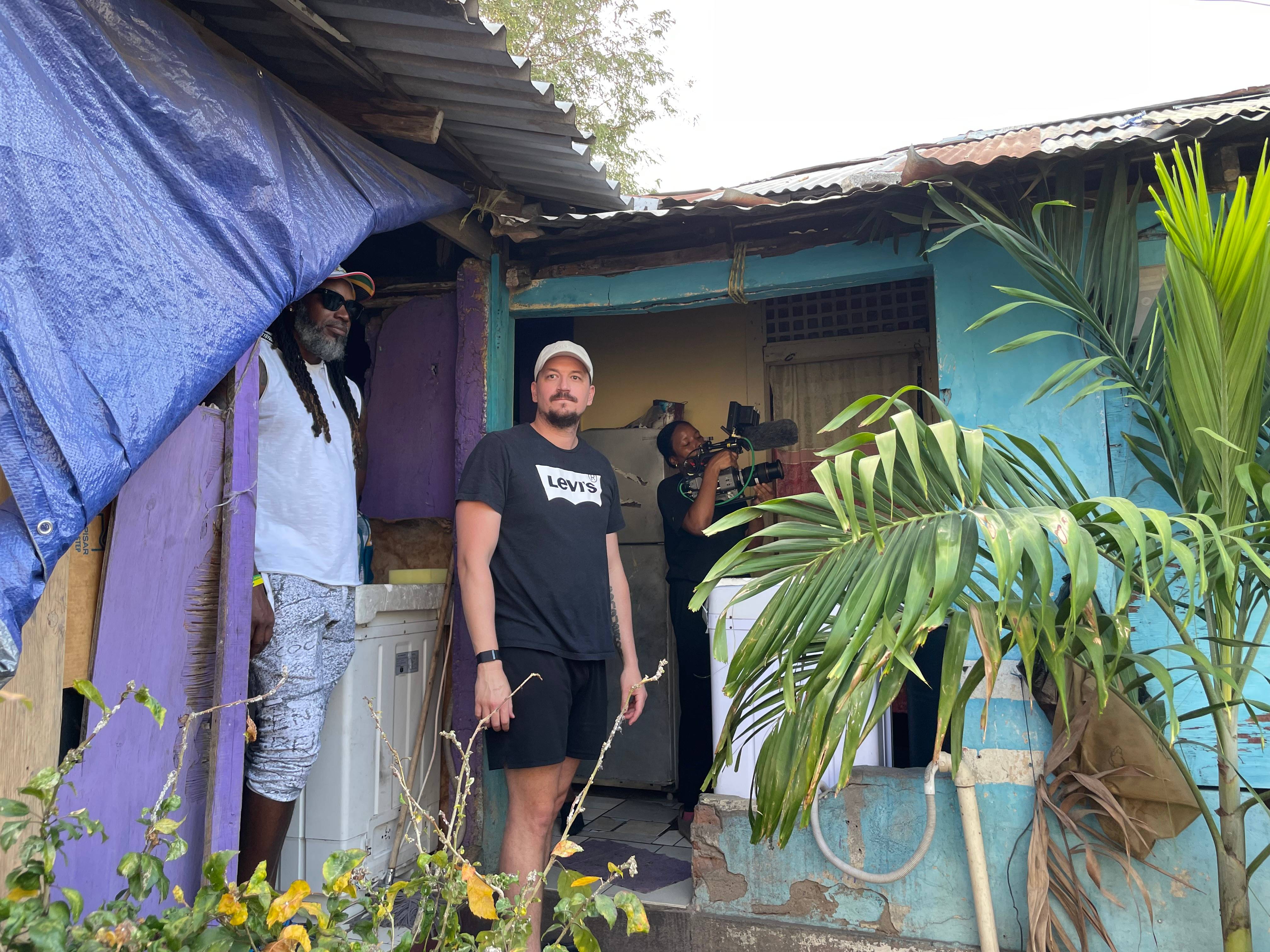 Sean Bodden on location with a camera operator and crew outside a weathered timber house.