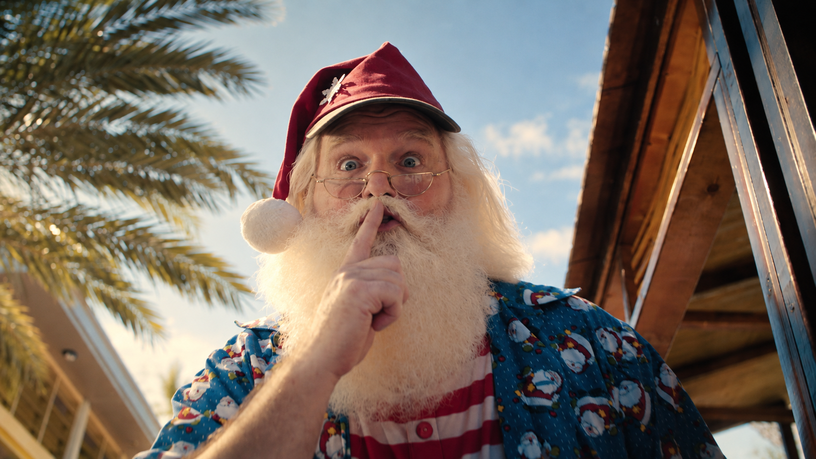 Santa, in a cap and holiday shirt, holds a finger to his lips beneath a palm tree.