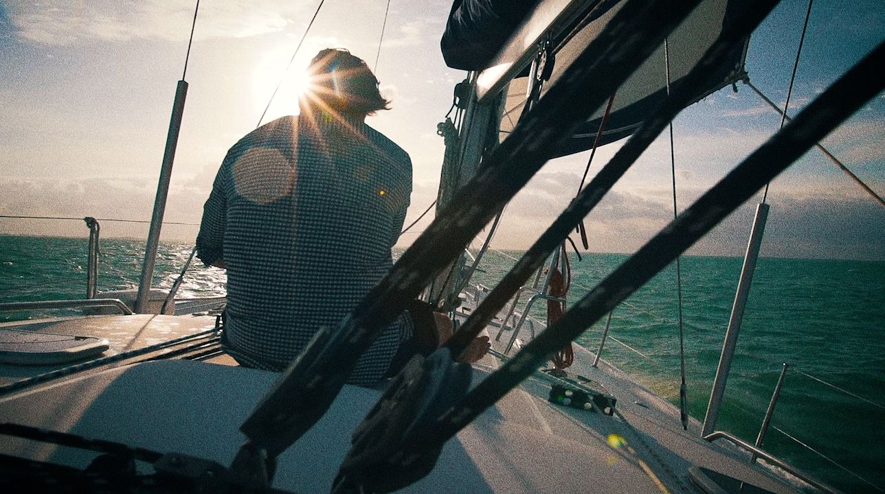 A man sits on the bow of a sailboat, sun flaring through the rigging.