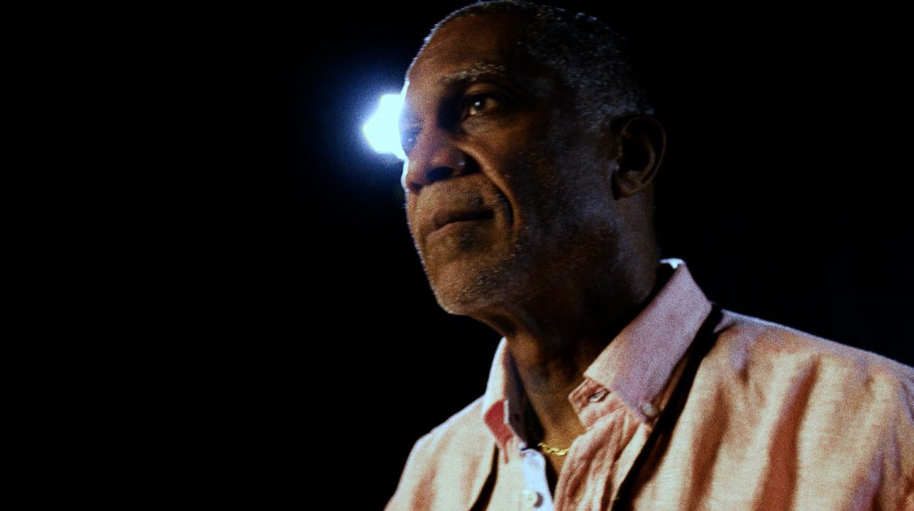 An older man in a pink shirt, lit by a single hard light.