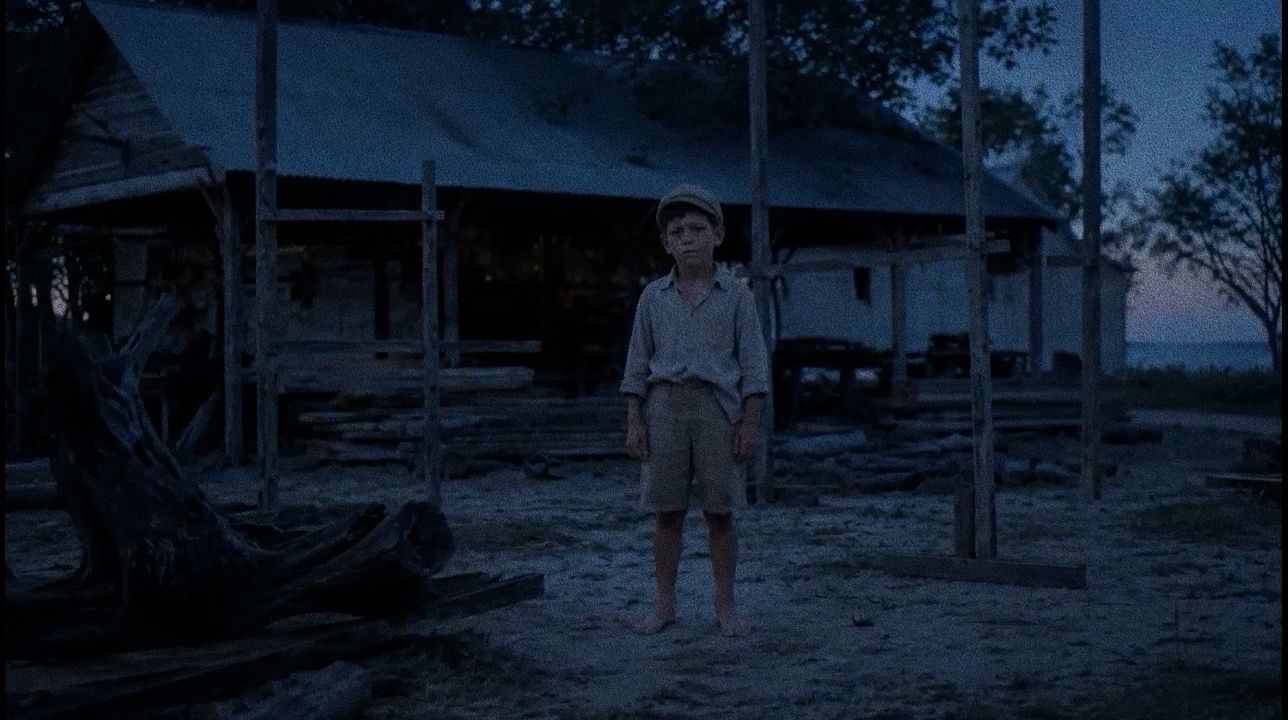 A boy stands barefoot in a dusk-lit boatyard.