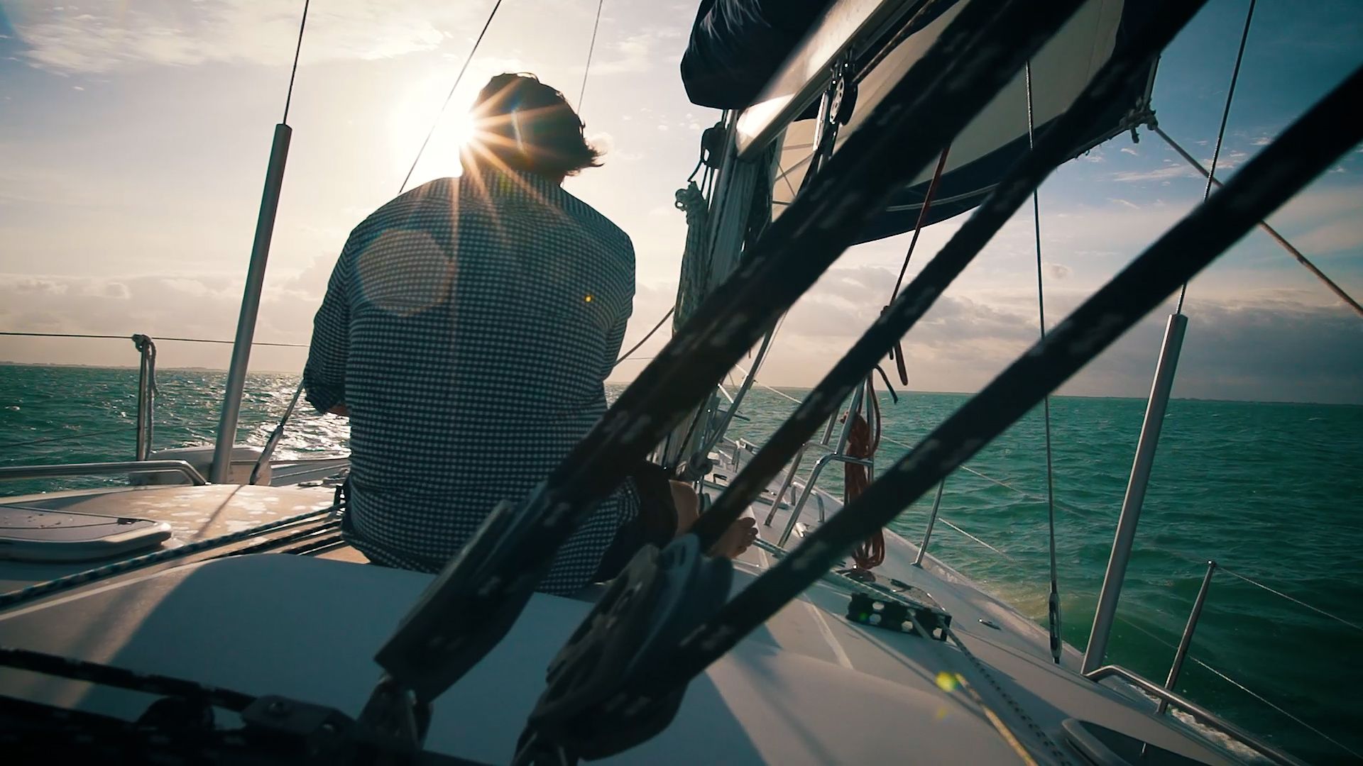 A man sits on the bow of a sailboat, sun flaring through the rigging.